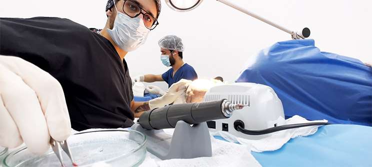 A doctor wearing a mask, glasses, and black scrubs holding tweezers during a hair transplant procedure. In the background, another medical professional works on a patient, highlighting teamwork in a clinical setting.