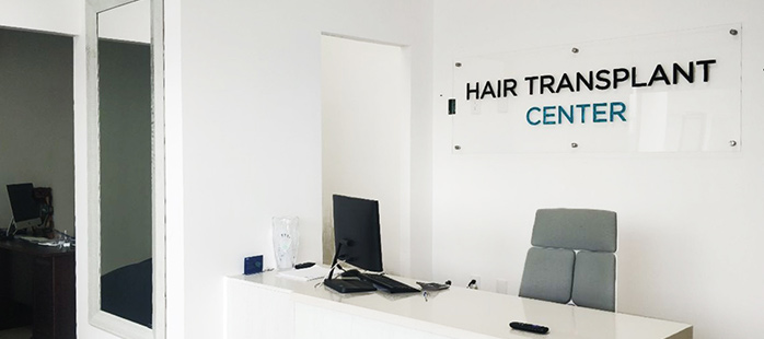 This image depicts the reception area of a hair transplant center, featuring a clean and minimalist design. A white desk with a computer monitor, phone, and other accessories sits in front of a modern acrylic sign that reads "Hair Transplant Center." The light gray chair and uncluttered space emphasize professionalism and welcoming aesthetics.