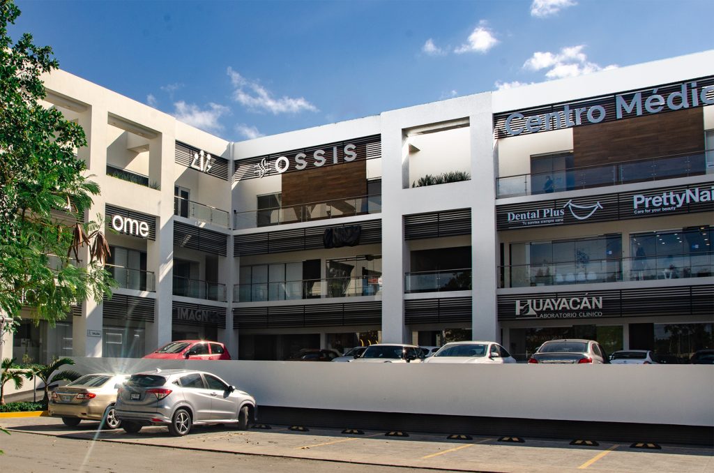 This image features the exterior of a modern medical complex housing a hair transplant clinic. The building is multi-storied with sleek architectural elements, including glass windows, clean white walls, and prominent signage for various businesses, including the clinic. The clear blue sky and parked cars add to the welcoming and professional atmosphere.