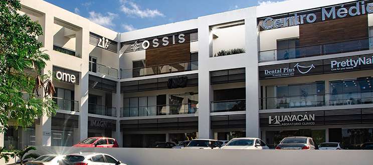 This image features the exterior of a modern medical complex housing a hair transplant clinic. The building is multi-storied with sleek architectural elements, including glass windows, clean white walls, and prominent signage for various businesses, including the clinic. The clear blue sky and parked cars add to the welcoming and professional atmosphere.