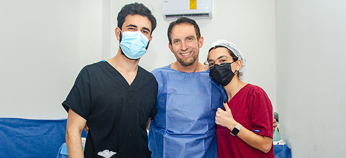 This image features two medical professionals posing with a patient dressed in a blue surgical cape. The healthcare workers, wearing scrubs and face masks, stand on either side of the patient, who smiles warmly at the camera.