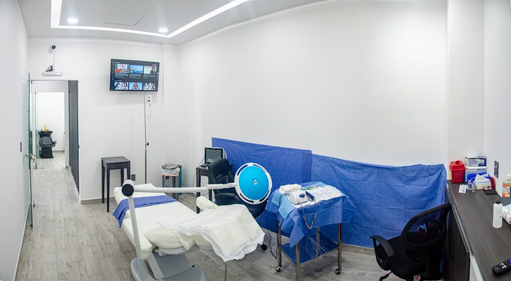 This image shows a modern and well-equipped medical room designed for hair transplant procedures. The space features a reclining chair, a surgical instrument table covered with blue sterile drapes, and advanced medical equipment, including monitors and lights. A wall-mounted screen and clean, minimalist design emphasize a professional and hygienic clinical environment.