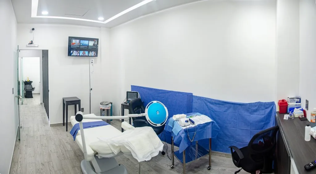 This image shows a modern and well-equipped medical room designed for hair transplant procedures. The space features a reclining chair, a surgical instrument table covered with blue sterile drapes, and advanced medical equipment, including monitors and lights. A wall-mounted screen and clean, minimalist design emphasize a professional and hygienic clinical environment.
