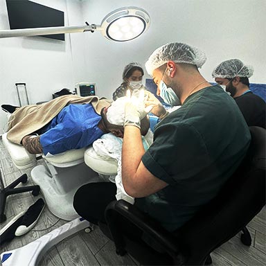 This image captures a hair transplant procedure in progress, featuring a team of medical professionals in a well-equipped clinical setting. A patient lies on a specialized reclining chair while a doctor, wearing a surgical cap and gloves, works under a bright overhead light. Additional team members are seen in the background, assisting with the procedure, emphasizing precision and teamwork in hair restoration treatments.