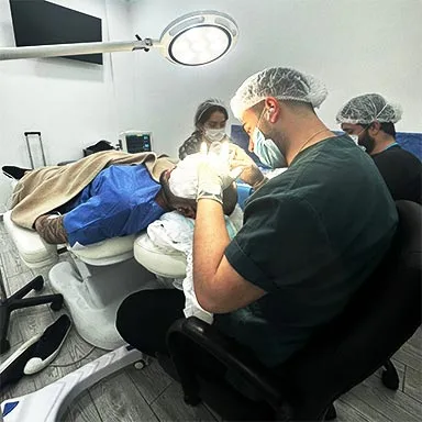 This image captures a hair transplant procedure in progress, featuring a team of medical professionals in a well-equipped clinical setting. A patient lies on a specialized reclining chair while a doctor, wearing a surgical cap and gloves, works under a bright overhead light. Additional team members are seen in the background, assisting with the procedure, emphasizing precision and teamwork in hair restoration treatments.