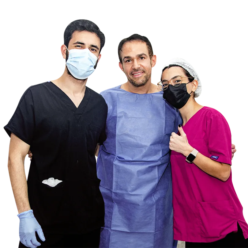 This image features two medical professionals posing with a patient dressed in a blue surgical cape. The healthcare workers, wearing scrubs and face masks, stand on either side of the patient, who smiles warmly at the camera. The image, set against a transparent background, conveys a sense of professionalism and care before a medical procedure.