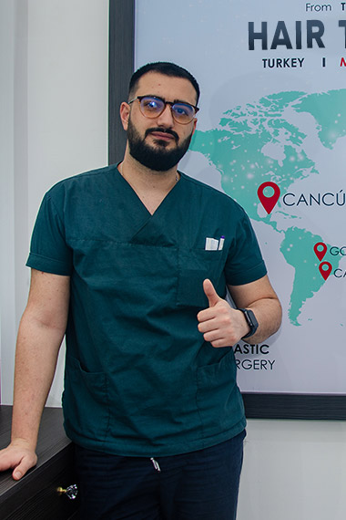 A person wearing teal medical scrubs gives a thumbs-up in front of a world map poster highlighting hair transplant surgery locations in Turkey, Cancún, and Costa Rica.