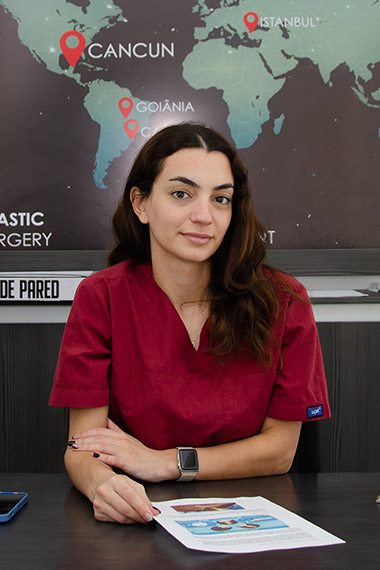 A woman in a red medical uniform sits at a desk with her hands resting on a document that features images and text. She has a calm and professional expression, wearing a smartwatch and a bracelet. Behind her is a world map with red location pins marking Cancun, Goiânia, and Istanbul, with partial text visible related to plastic surgery.